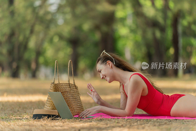 美丽的白人女性性感的女人在红色泳装比基尼戴着太阳镜躺在地上工作在她的电脑笔记本电脑放松在树林里的海滩