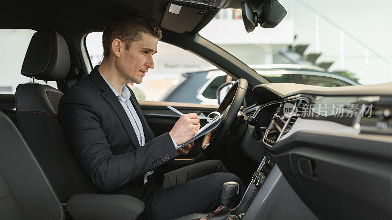 年轻的汽车销售员坐在全新的汽车里，在现代化的经销商，全景进行检查，记录数据