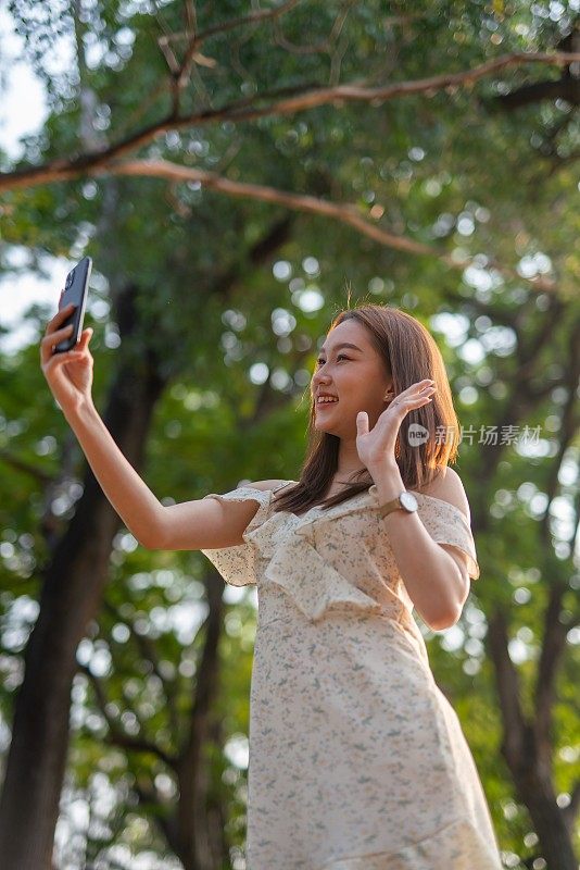 年轻的亚洲女子在公园用手机自拍。在郁郁葱葱的绿色森林里，美女视频通话