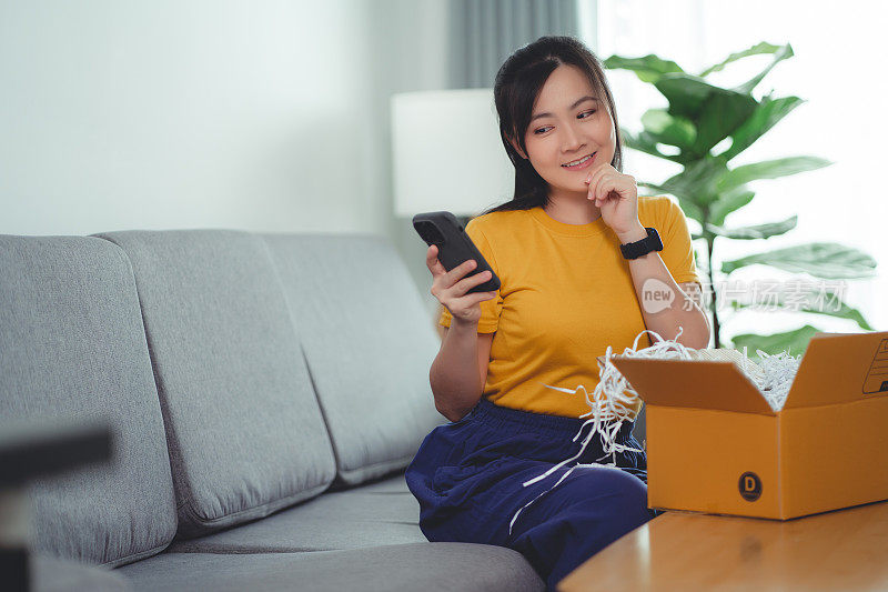 亚洲女人打开纸箱，开心兴奋地坐在家里客厅的沙发上玩智能手机。