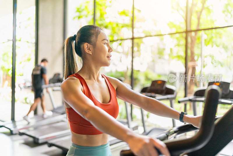 一名亚洲女性在光线充足的健身房里骑着健身车，并拥有花园景观