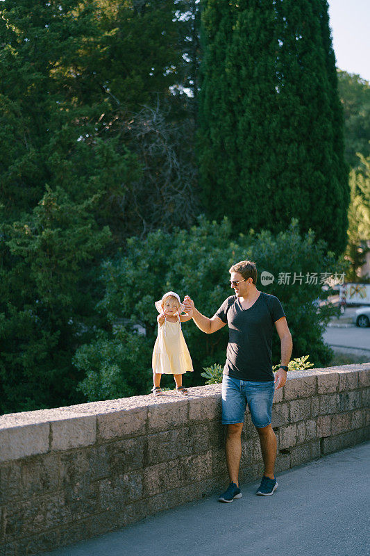 爸爸牵着一个小女孩的手沿着公园里的石栅栏走