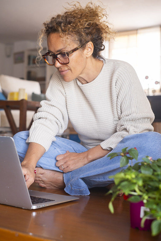 一位中年女士坐在地板上舒舒服服地使用笔记本电脑独自在室内科技无线休闲活动。现代女性在电脑上工作。女人浏览网页和写作