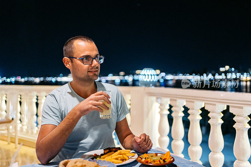 在埃及卢克索，一名年轻男子在餐厅的阳台上享用晚餐，窗外是尼罗河的夜景