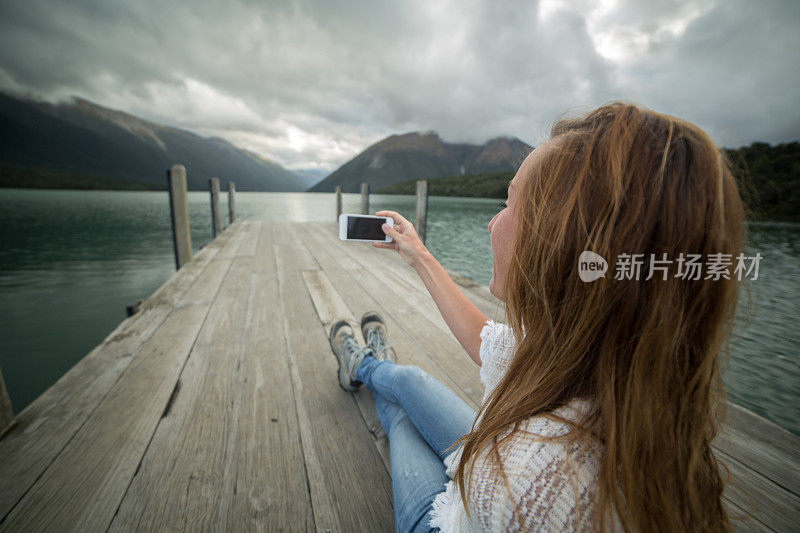 年轻的女性坐在湖边的码头上使用手机