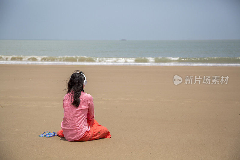 女孩坐在海滩上，听着音乐，面朝大海