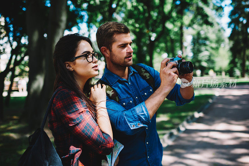 男男女女在公园散步，在旅行中用老式相机拍很酷的照片。相爱的情侣背着背包，穿着休闲装，一起站在户外拍照