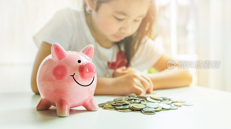 一个可爱的粉红色储蓄罐和一堆硬币在桌子上的背景是亚洲小女孩