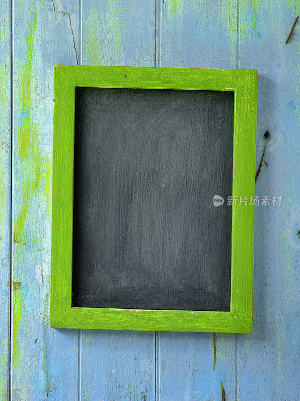 一块古老的风化的绿色木框架黑板挂在一个古老的风化的蓝色和绿色木面板墙的背景。