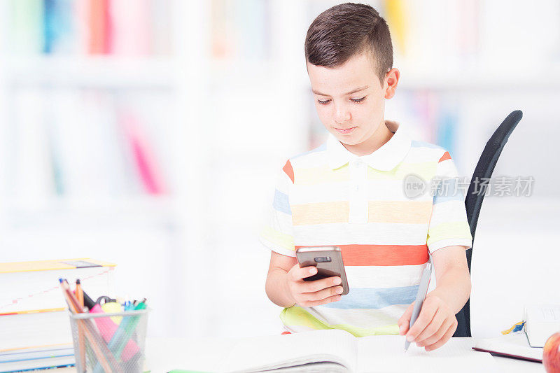 可爱快乐的男孩用手机做家庭作业。回到学校的概念。滑稽情绪少年坐在课桌前写作业。书架在背景中失去焦点