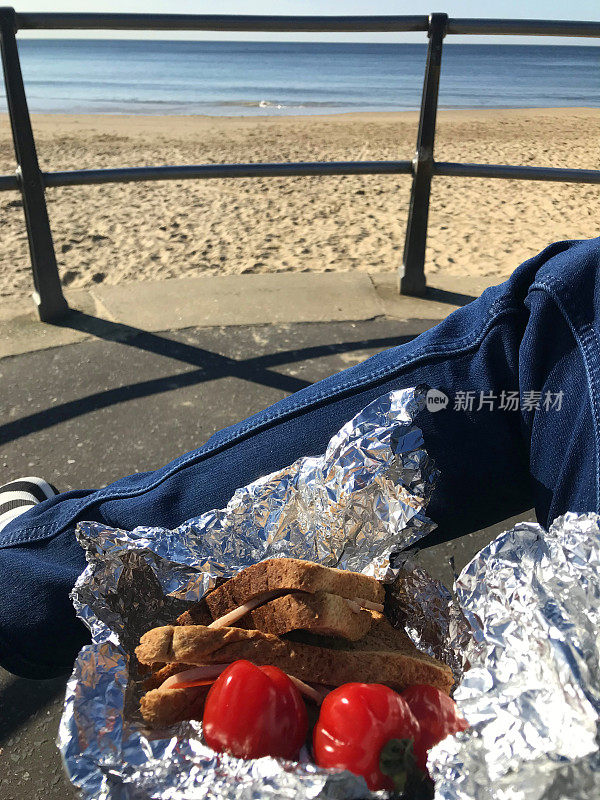 一个男人坐在长椅上，在暑假吃英国野餐在海滩散步与棕色切全麦面包火腿三明治包裹在铝厨房箔樱桃李子西红柿和塑料瓶水，海边野餐穿着蓝色牛仔裤