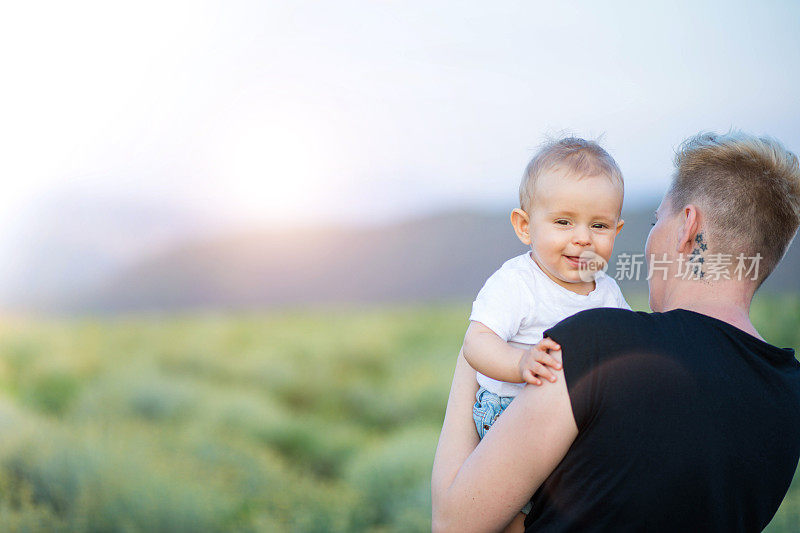一个年轻的母亲背着她的婴儿男孩，而一起走在草地上的自然黄花。婴儿正从她妈妈的肩膀后面看过去
