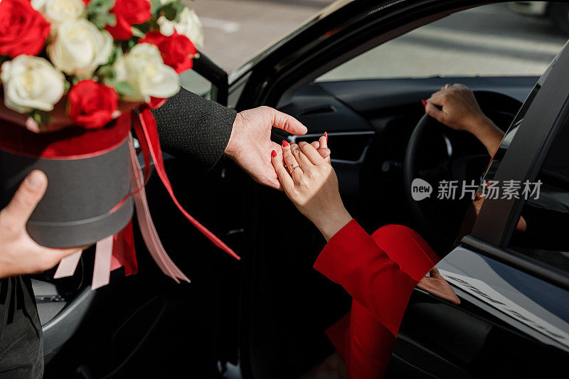 一名彬彬有礼的男子手持一束鲜花，帮助身着红色套装的女商人下车