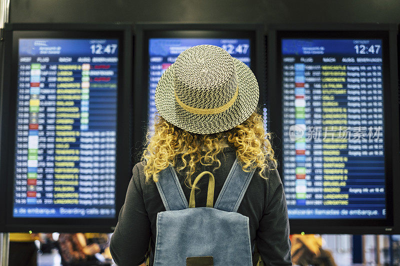 在机场或火车站旅行的人概念-从一个金发卷曲的女性女人的背影看和检查时间出发或到达的屏幕上的蓝色背包