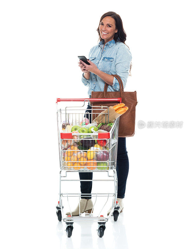 白人女性站在白色背景前，穿着裤子，拿着购物车，用着智能手机