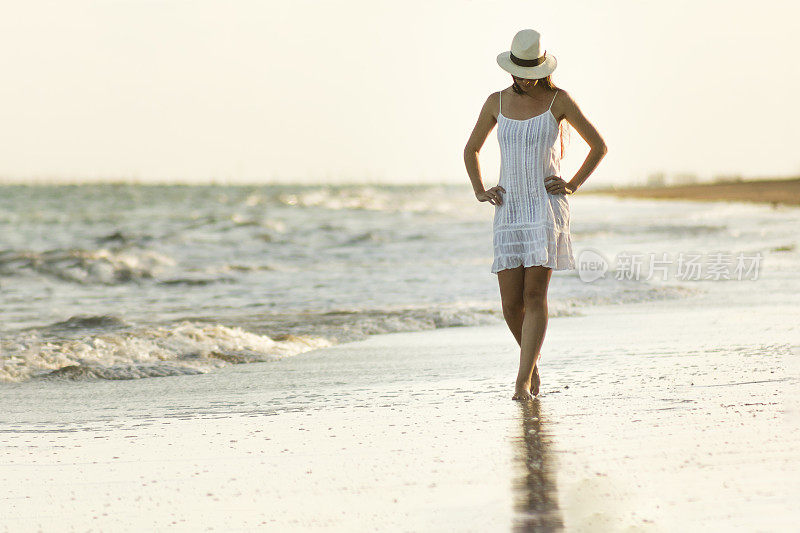在巴西东北部的阿拉格斯，美丽的女人走在日落的海滩上的肖像。她穿着白色的衣服，戴着太阳镜和草帽