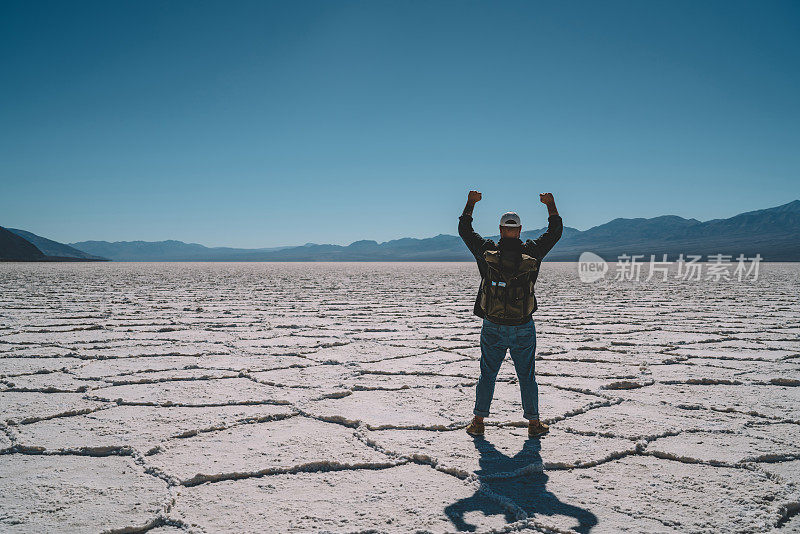 在badwater盆地的沙漠土地上，一个人在积极的旅行中举起双手庆祝成就的背影，年轻的潮男在荒野沙漠中完成徒步旅行，感觉成功和自由