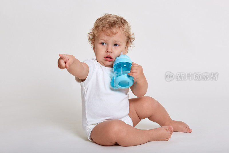 小男孩独自坐在地板上喝水，白色背景下，看着并指向别处，穿着紧身衣，有着金色的卷发，孩子戴着蓝色的婴儿罩杯。