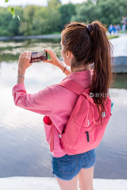 夏天，一名女子在城市的湖边、河边池塘边，用智能手机拍摄风景，从背后观看，背后背着一个粉色背包，在互联网上录制视频，应用智能手机摄像头。