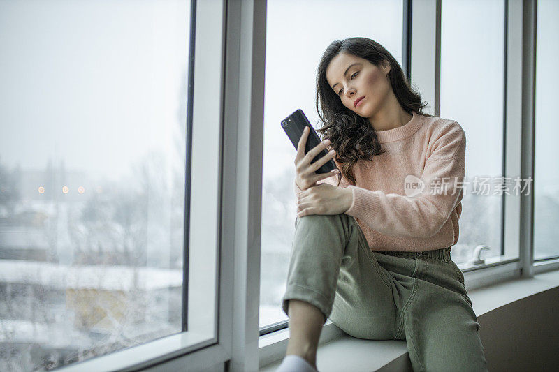 抑郁的女人在家里感到难过。女人悲伤地看着窗外。一个沮丧孤独的女人独自站在那里，一边打电话一边看窗外。