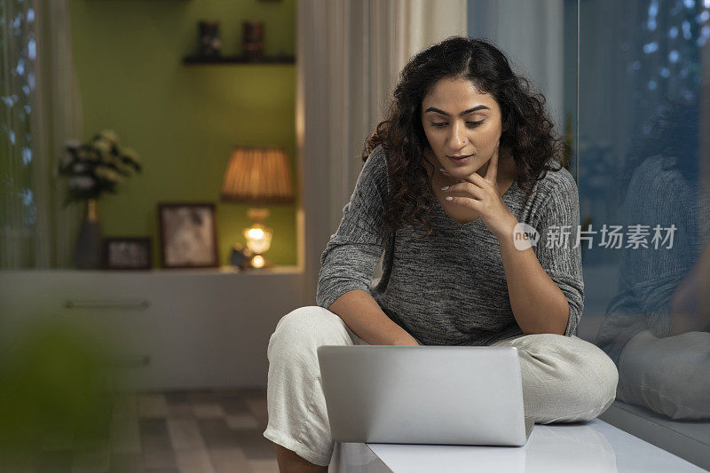 一个年轻女性坐在家里窗台上使用笔记本电脑的照片，库存照片