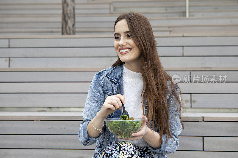 一名年轻女子穿着花裙子、白色套头衫和牛仔夹克，坐在木质台阶上，从一个圆形塑料(聚对苯二甲酸乙二醇酯)容器里吃着沙拉