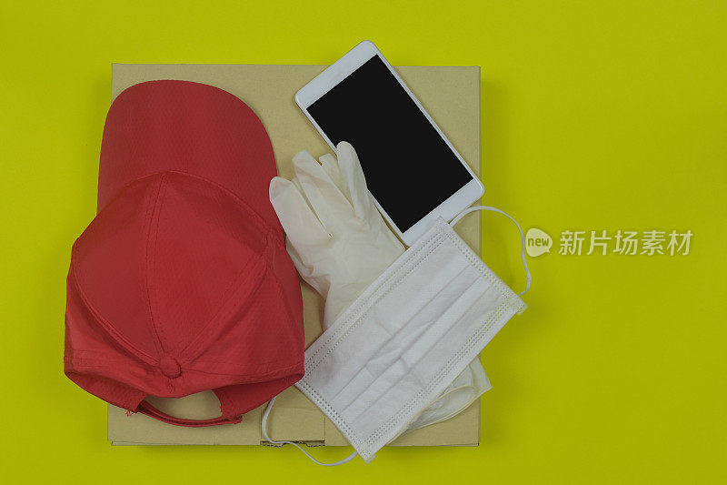披萨外卖的概念。黄色背景上有手机、帽子、医用橡胶手套和口罩的披萨盒。