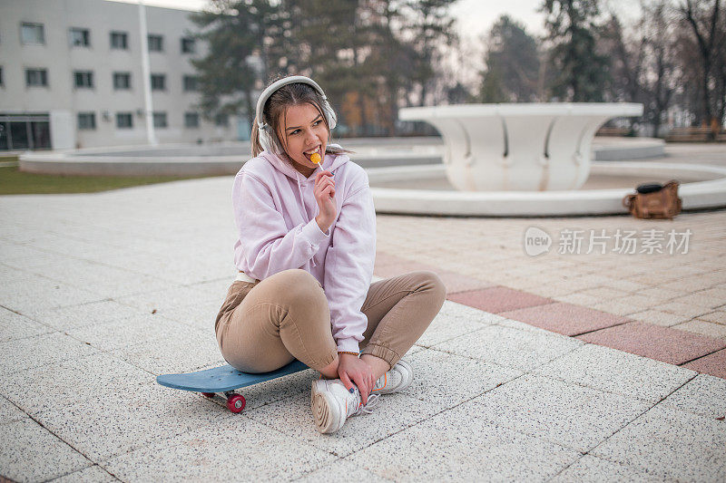 年轻女子一边舔着棒棒糖一边坐在滑板上听着音乐