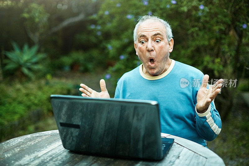 一位老人坐在郁郁葱葱的花园里的笔记本电脑前，看起来很震惊和恐惧