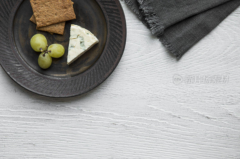 四份D'Ambert奶酪放在盘子里，还有饼干和葡萄，一张餐巾纸放在白色的木桌上——从正上方可以看到