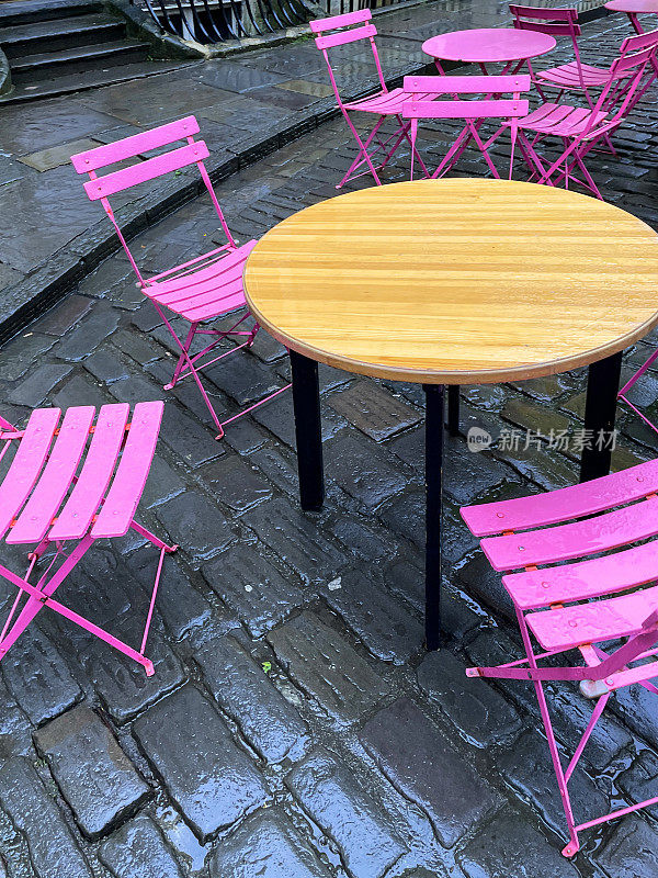 色彩鲜艳的图像，圆形圆形金属庭院桌椅涂成亮粉色，露天就餐在鹅卵石街道上，重点在前景