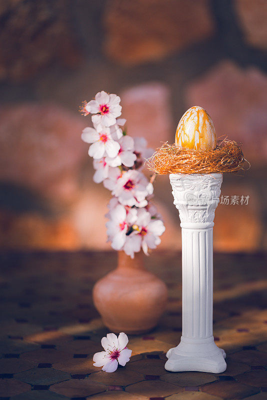 一个金色的复活节彩蛋在一个古老的乡村桌子上的白色柱子上的金色鸟巢里，杏仁花在阳光和阴影的结合中