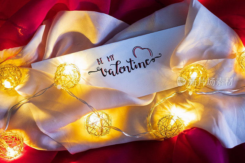 情人节可爱的便条写在白纸上，上面覆盖着节日灯链球、花环和粉色台布，一个装饰的设计想法