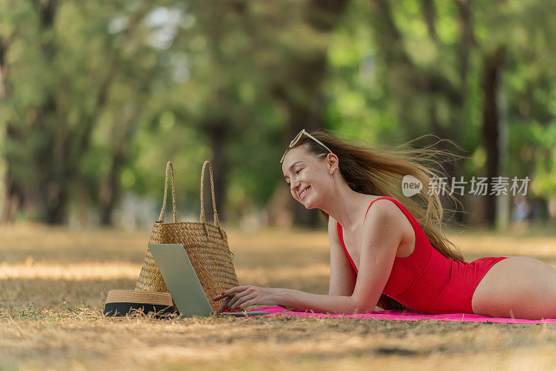 美丽的白人女性性感的女人在红色泳装比基尼戴着太阳镜躺在地上工作在她的电脑笔记本电脑放松在树林里的海滩