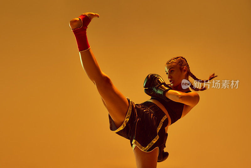 强有力的腿。综合格斗选手，十几岁的女孩训练，在霓虹灯的橙色工作室背景下用腿踢