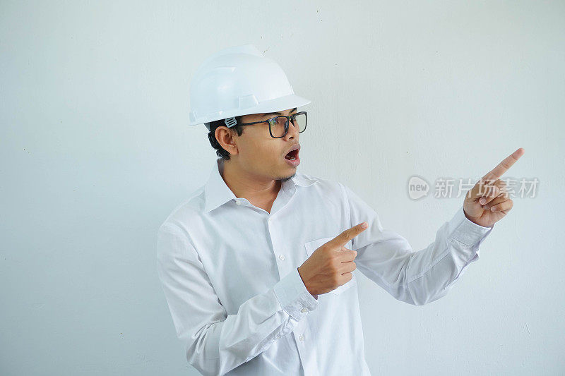 震惊的年轻亚洲建筑师男子戴着白色安全帽，手指指向左侧，孤立在白色背景上