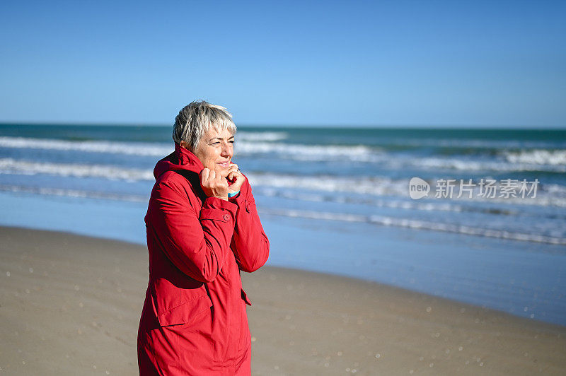 一个头发灰白的女人在刮风的天气里独自走在沙滩上的画像。