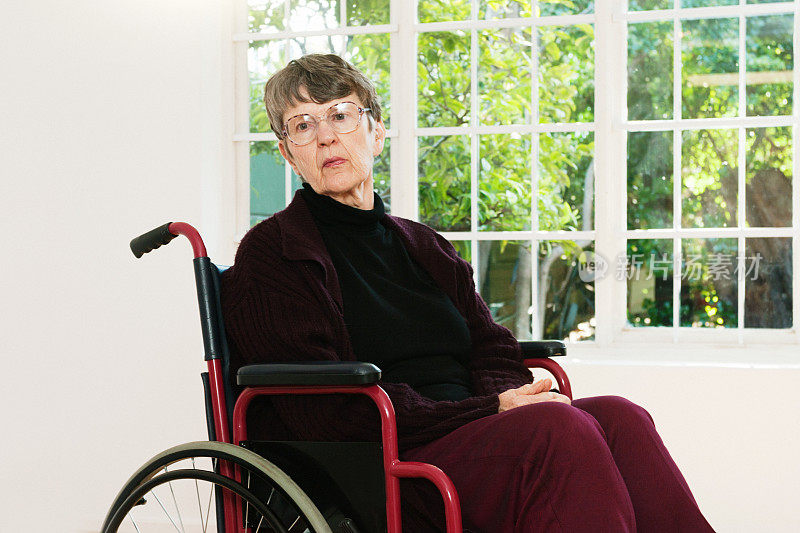 一位70多岁的老妇人坐在轮椅上，靠着花园明亮的窗户，严肃地看着镜头