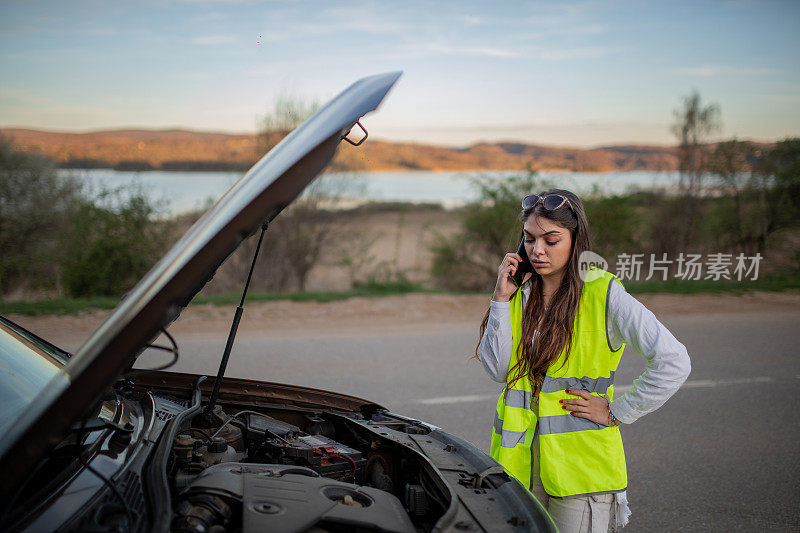 年轻的压力很大的女子站在她抛锚的汽车旁边，用手机求救