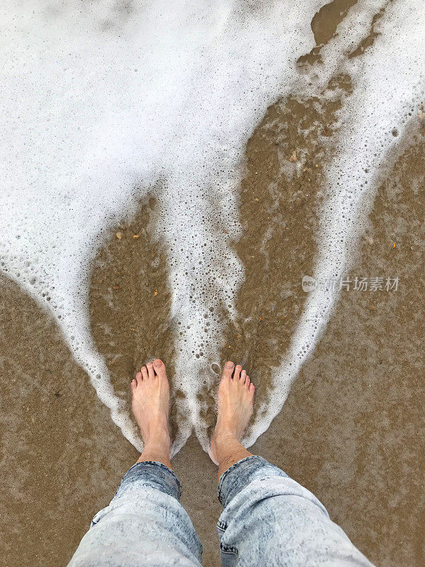 光着脚的男人两只白脚站在沙滩上划着海浪，在退潮时溅起水花，光着脚陷在正在下沉的湿沙子里，留下脚印，海水的水滴和湿牛仔裤被水溅起，沙子被冲走，溅起泡沫