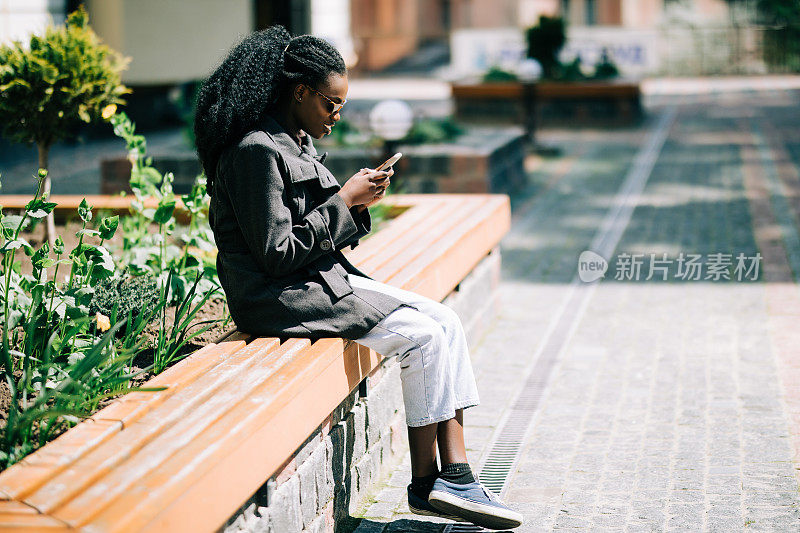 一个快乐的年轻非洲妇女坐在外面的长凳上看手机短信的肖像