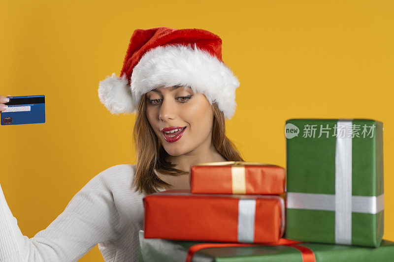 性感的年轻成年女人戴着圣诞帽在圣诞节看圣诞礼物包装的圣诞纸和显示她的信用卡