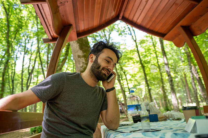 黑胡子独自英俊快乐的男人微笑，在森林野餐的一个阴凉下打电话在公共公园概念