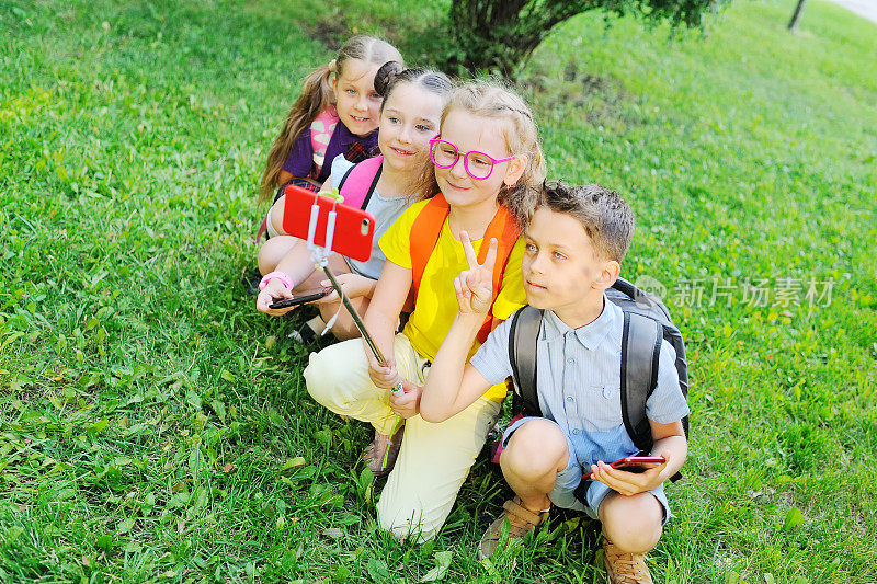 一群学生坐在草地上，穿着五颜六色的衣服，背着书包，微笑着玩耍，用自拍杆上的智能手机相机拍照。回到学校，电子产品，智能手机成瘾