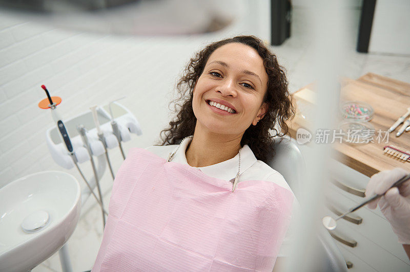 在现代牙科诊所，非裔美国美女带着美丽的牙齿微笑，坐在牙医椅子上等待牙科检查。口腔护理，口腔卫生概念
