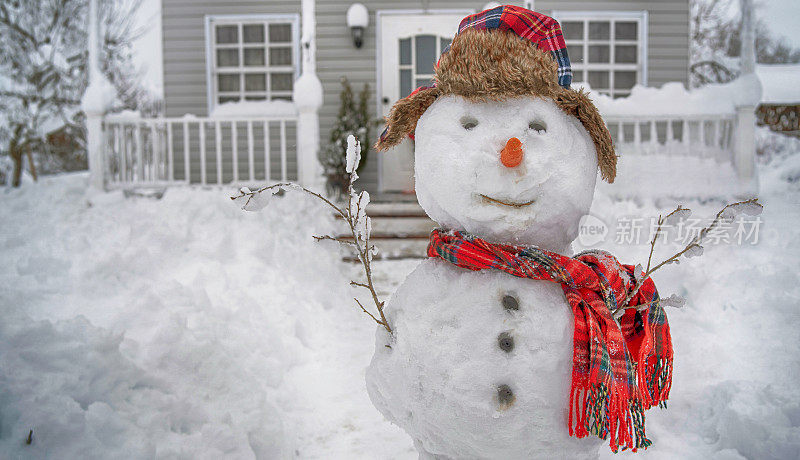在冬天的一天，一个可爱的微笑的雪人，戴着红色的围巾和帽子，站在下雪的门廊前