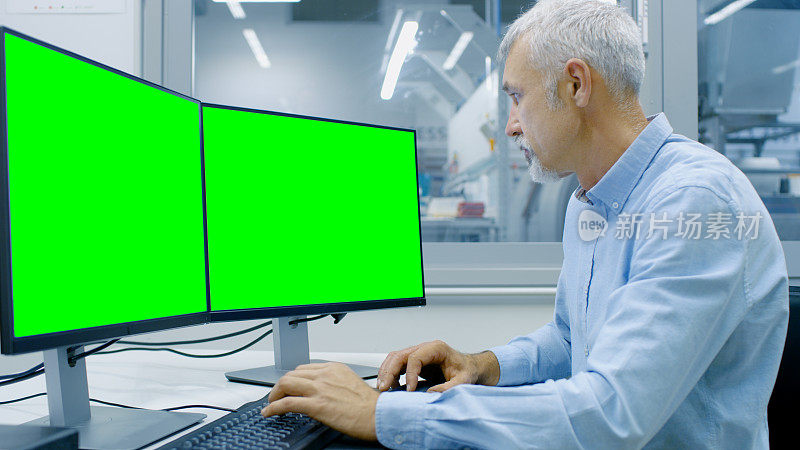 工程师工作在一个有两个显示器的计算机上，一个有色度键伟大的模型模板和CAD程序中的其他组件。可见办公室外窗口组件制造工厂。