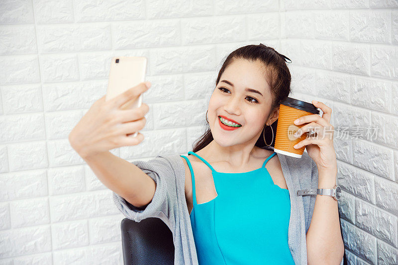 年轻的亚洲女性在咖啡馆用智能手机与咖啡杯自拍
