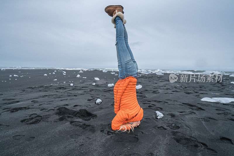 年轻女子头倒立瑜伽姿势在黑沙滩附近的冰和海在冰岛