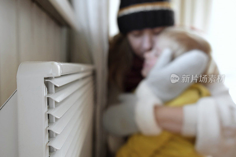 冻住了的母子穿着毛衣，冬天冷得发抖，抱着电暖炉。女孩和孩子在暖气片附近暖手。不舒服的呆在家里。天然气危机。